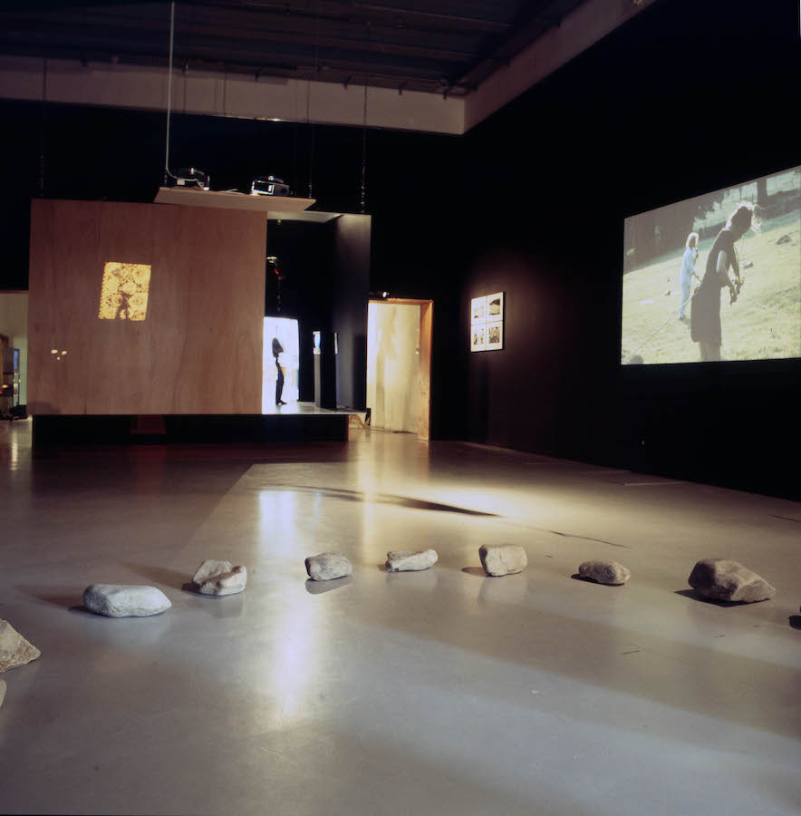 A room with rocks on the floor in the shape of a half circle, a video projection and photographs are on the wall to the right.