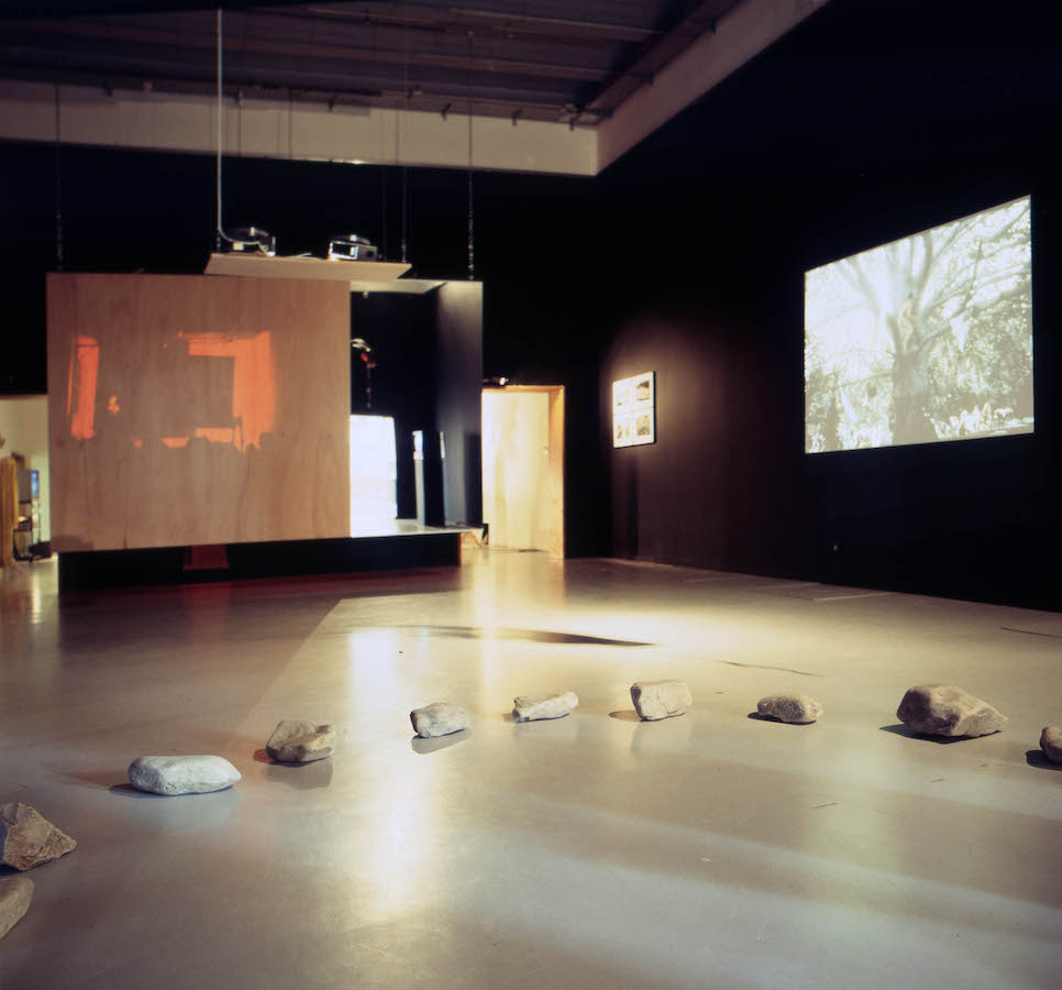 A room with rocks on the floor in the shape of a half circle, a video projection and photographs are on the wall to the right.