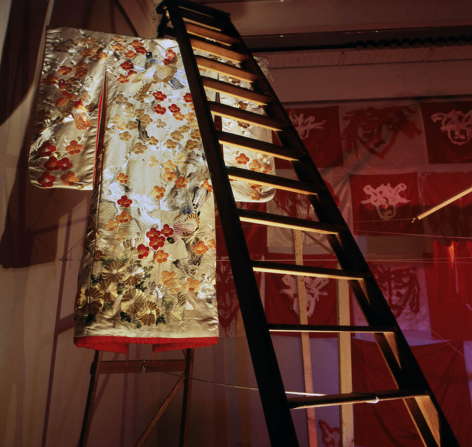 A robe hangs underneath a ladder, in the background is a wall with various paintings of heads.