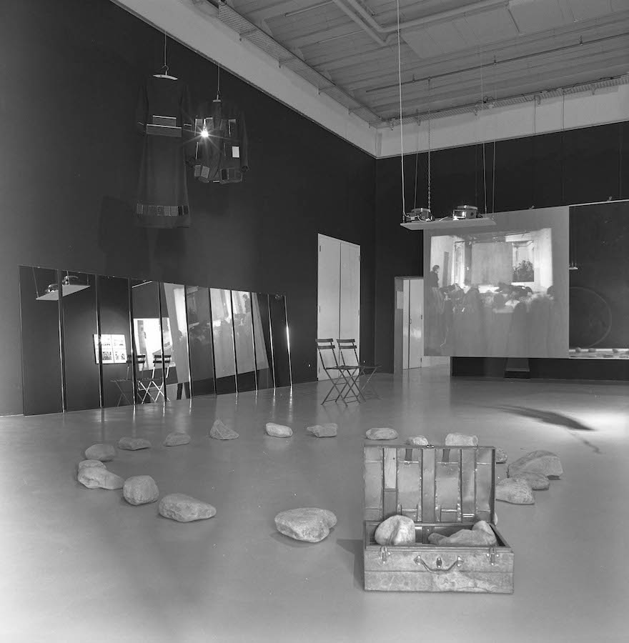 A room filled with various props including: rocks in the shape of a circle on the floor, a case full of rocks, full length mirrors along the wall, a dress and a jacket hanging from the ceiling and a video projection.