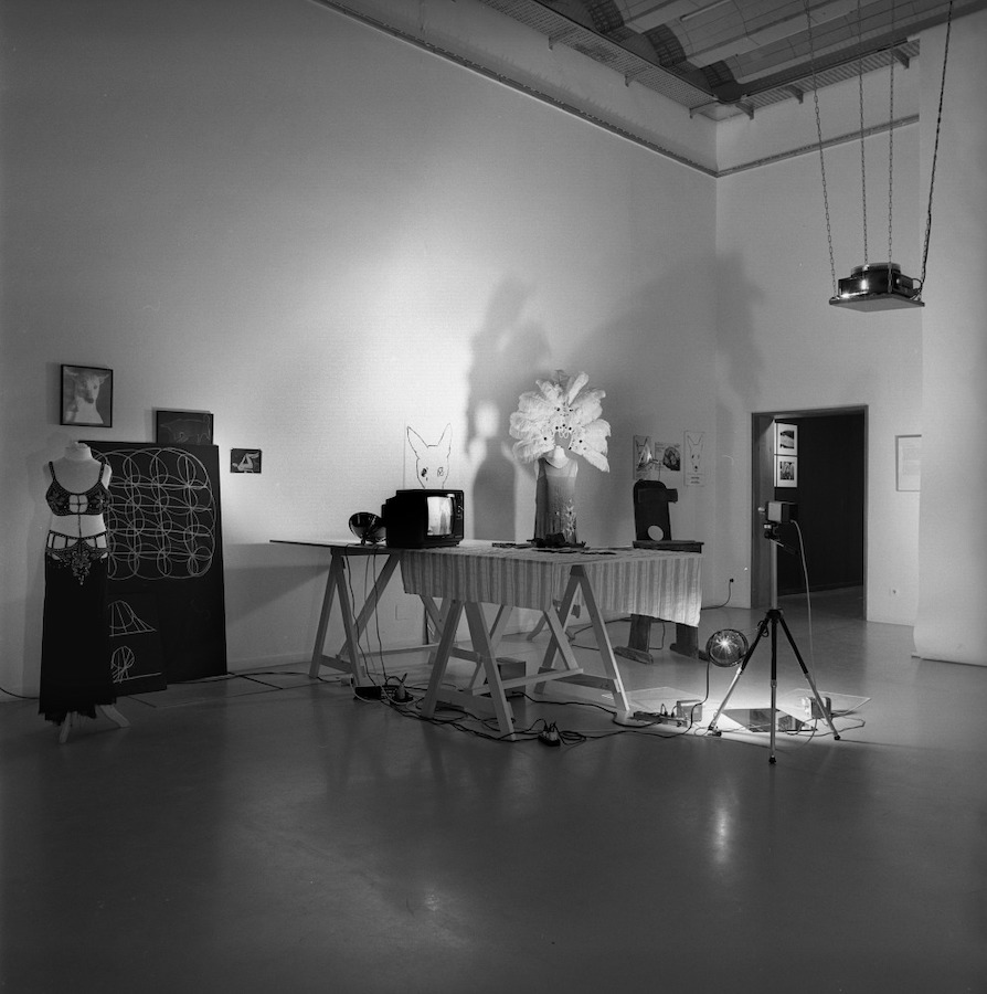 Various props placed in a room. In the center is a camera on a tripod facing a TV monitor on top of a table with a cloth draped over it. Behind the TV is a light illuminating a drawing of a dog’s head with four eyes hanging on the wall and Jonas’s dress and peacock-feathered headpiece placed on a mannequin. Another mannequin is located to the left of the TV that is wearing Jonas’s bra top and skirt; behind the mannequin is a drawing and various photographs hanging on the wall. There is a projector hanging from the ceiling.