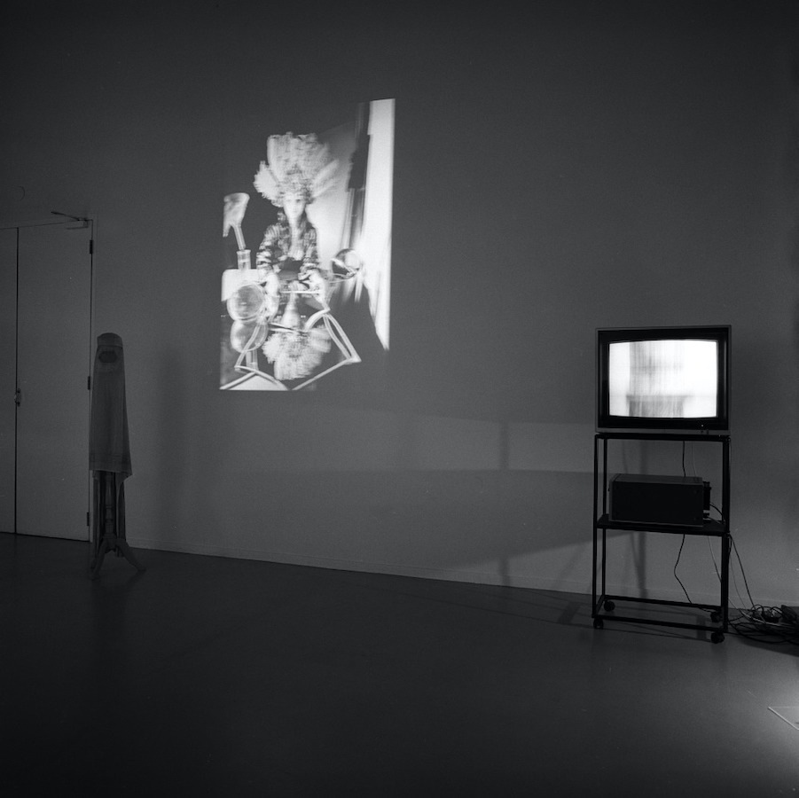 Jonas’s performance is projected onto a wall. To the left is an object standing on the floor. To the right is a TV monitor placed on a cart.