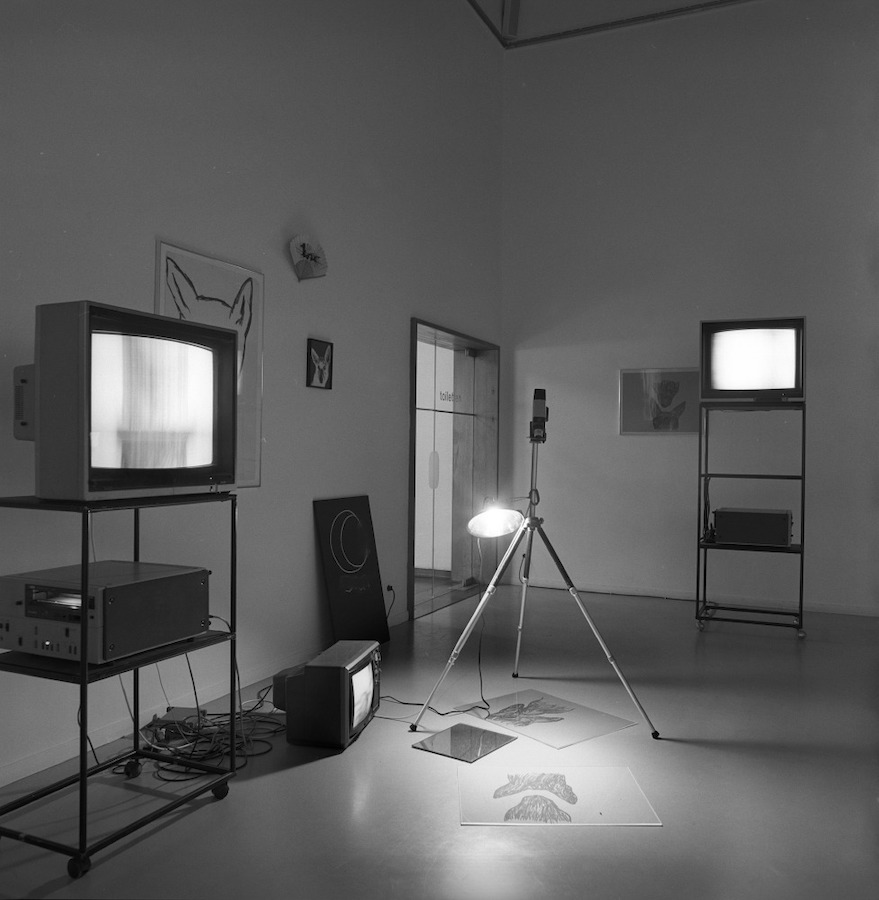 Various props placed in a room including: a camera on a tripod, two TV monitors on carts and various drawings hanging on the wall. On the floor there are drawings, a TV monitor, and a mirror which are illuminated by a light attached to the tripod.