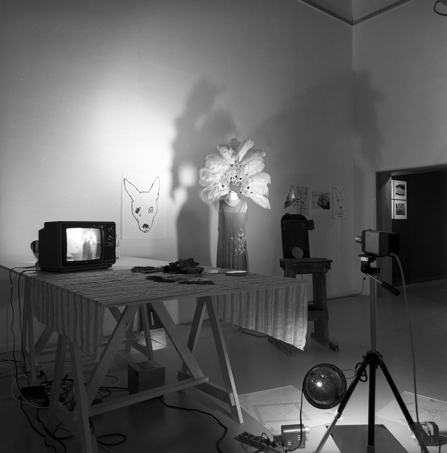 A camera on a tripod faces a mannequin wearing Jonas’s dress and peacock-feathered headpiece, and a drawing of a dog’s head with four eyes. There is a table with a TV monitor placed on top of a table with a cloth. On the floor is a mirror and various drawings.