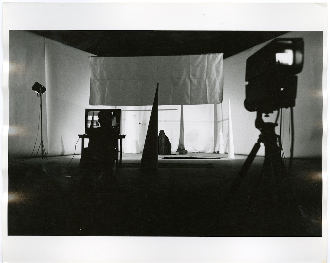 A camera on a tripod faces Jonas while she performs; there are various sized cones and a TV monitor placed on a table. There is a silhouette of a figure in front of the TV.