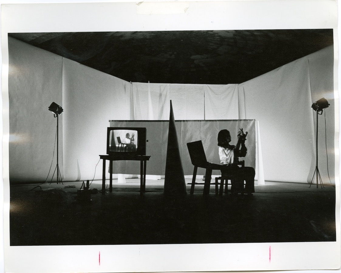 Jonas sits on a chair next to a child’s school desk with a lift-open top; there is a large cone and a TV monitor on a table that shows footage of Jonas.