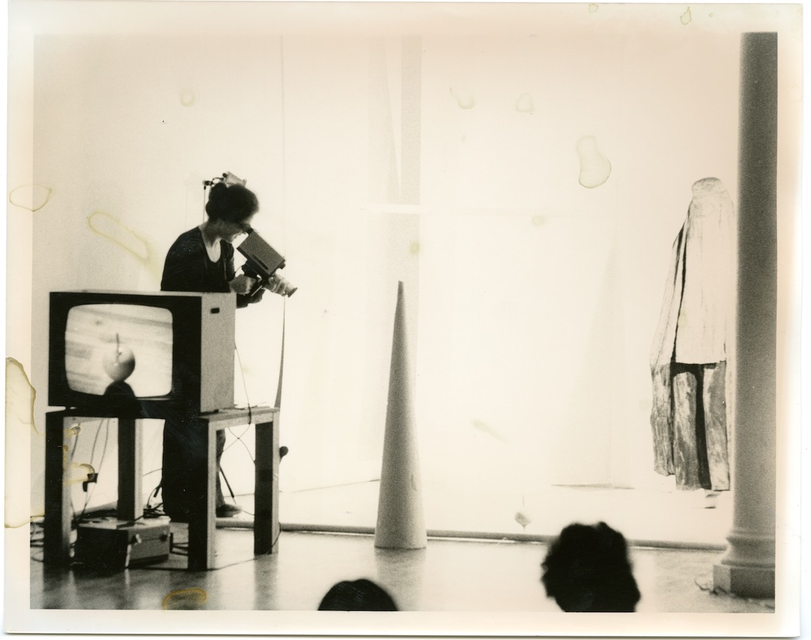 A camerawoman stands in front of a cone; to the left is a TV monitor on top of a table and to the right is a performer wearing a chadri. Audience members can be seen in the lower right corner.