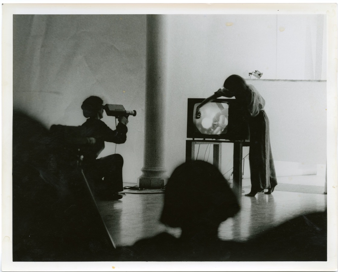 View from the audience, a camerawoman films Jonas as she interacts with a TV monitor on top of a table.