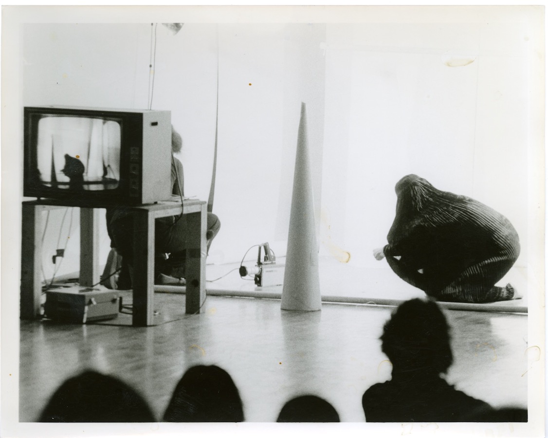 View from the audience, a camerawoman films Jonas performing. There is a cone and a TV monitor placed on a table projecting the film to the audience.