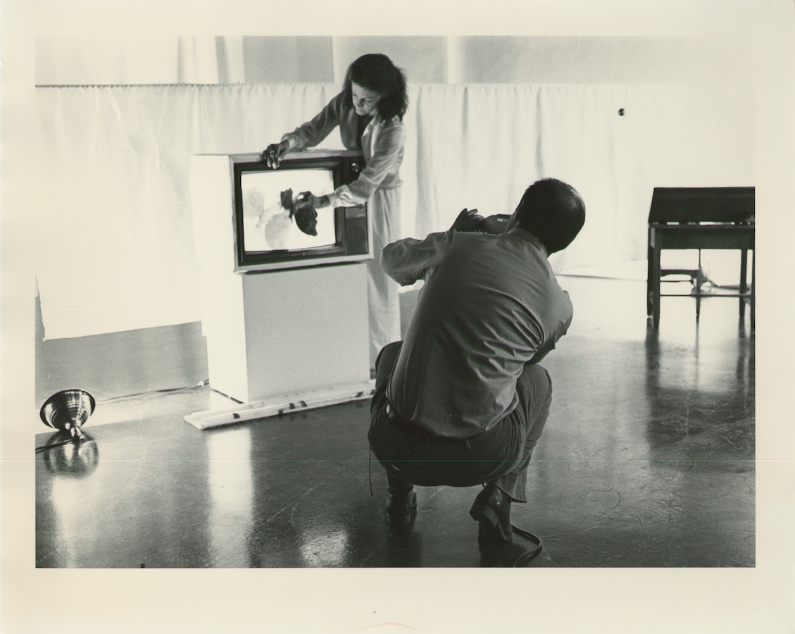 A cameraman films Jonas as she stands next to a TV monitor on a stand and holds a plastic butterfly in front of the monitor’s screen, which displays footage of the same butterfly.