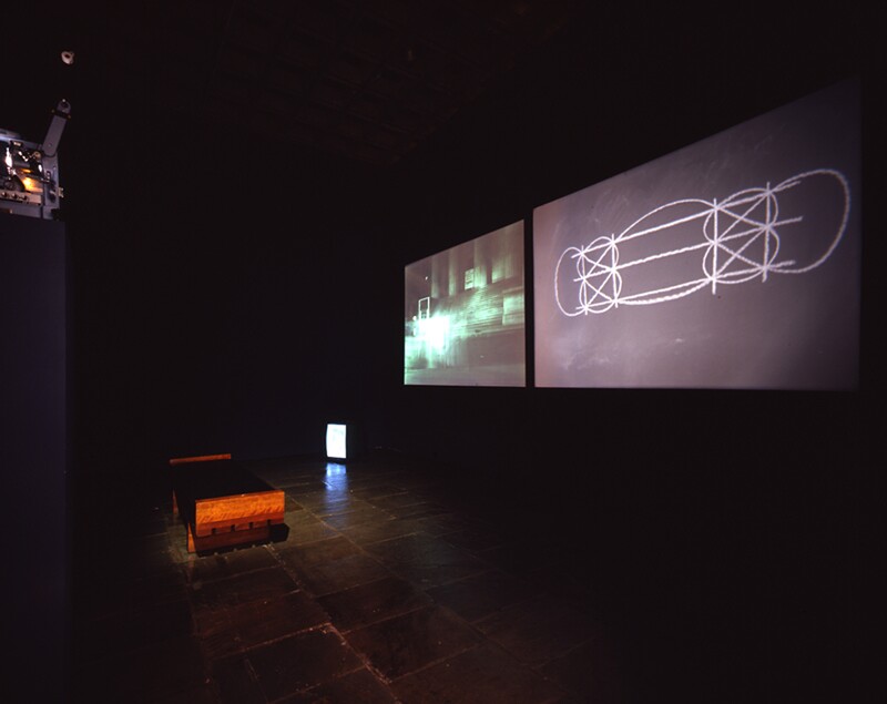 Two projections of Jonas’s performance are on the wall in front of a bench. In the corner is a TV monitor on the floor.