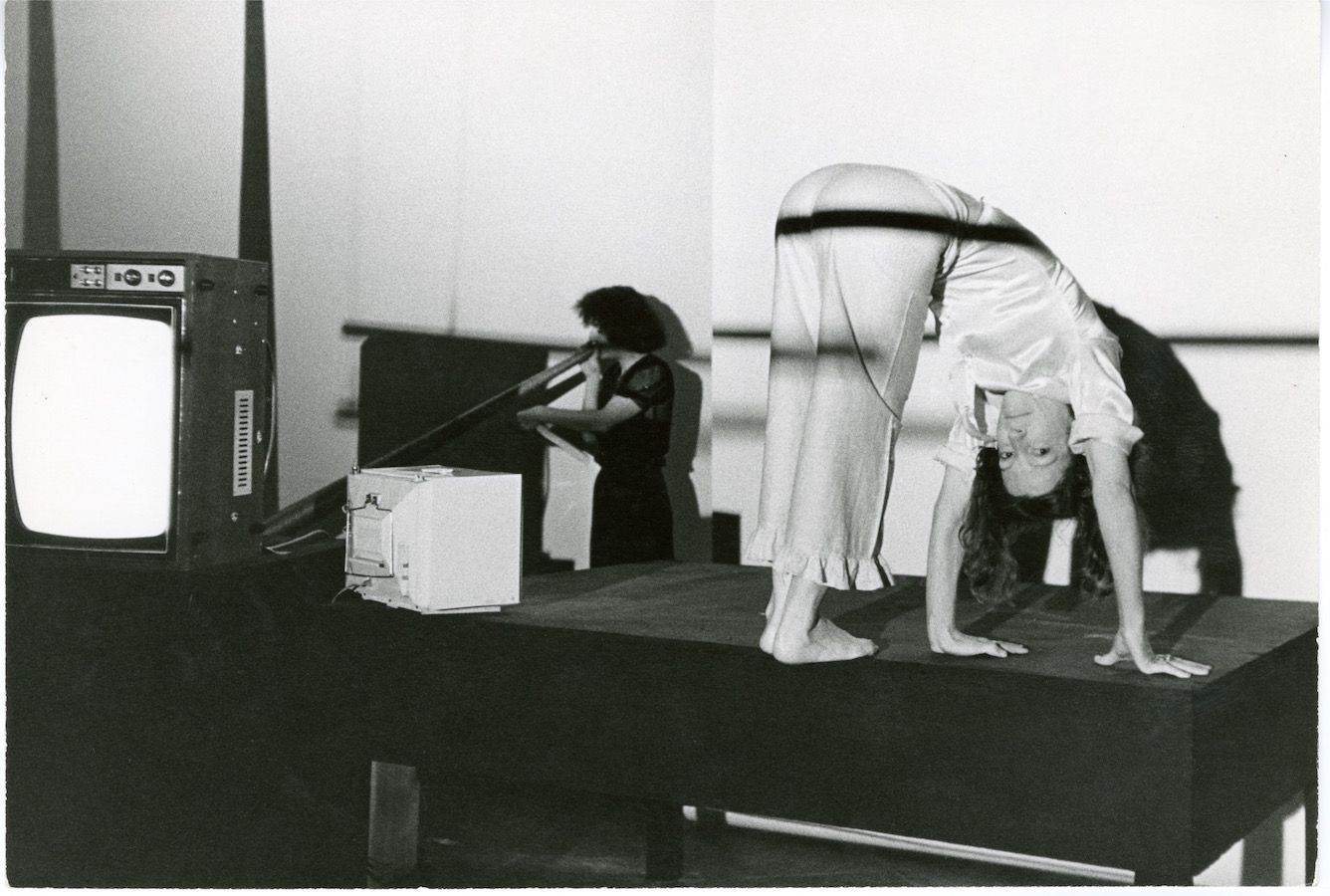 Jonas staring at the camera while bending over on top of a stage. There are two TV monitors, one on the stage and one on a stand, and another performer in the background with her mouth to a large cone.