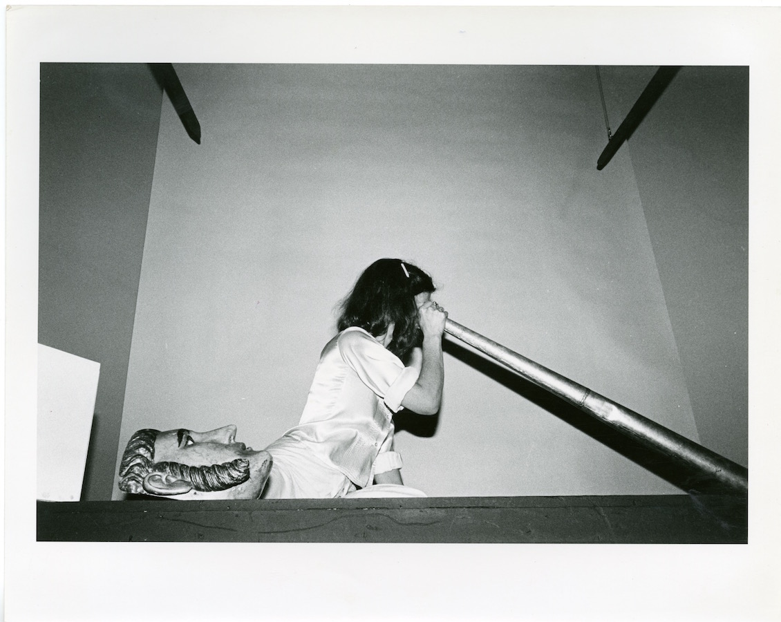 Jonas sitting on top of a stage looking down a large cone, next to her is a mask