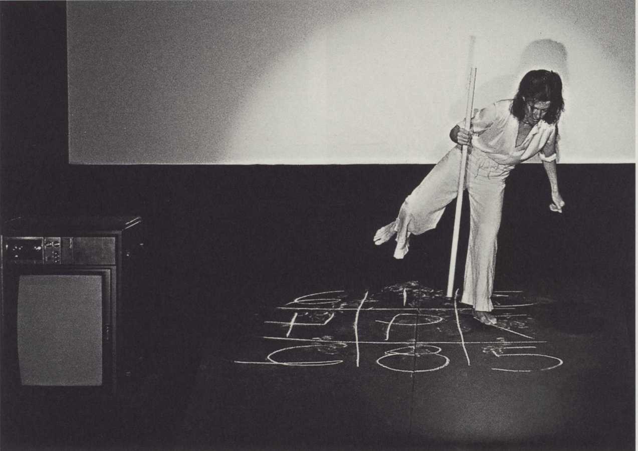 Jonas standing on top of a chalk drawing on one foot and holding a long stick. To the left is a TV monitor.