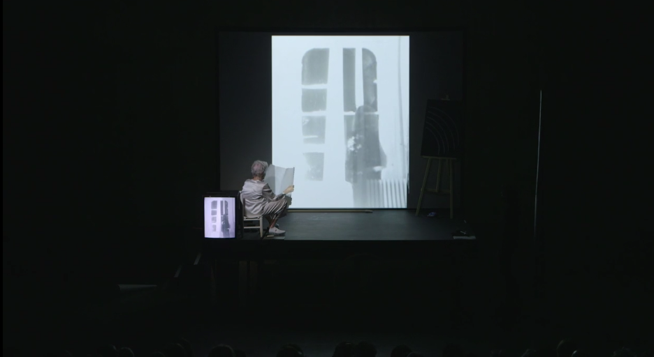 Jonas on stage sitting in a chair facing towards a projection of a window, holding a piece of paper. There is a chalk drawing placed on an easel to the right and a TV monitor to the left.