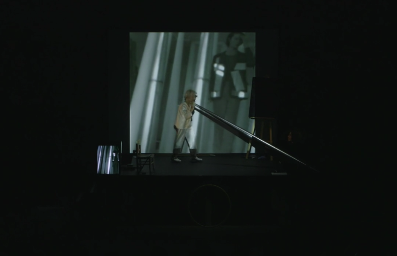 Jonas on stage in front of a projection, holding a large cone. There is a chalk drawing placed on an easel to the right and a TV monitor to the left.