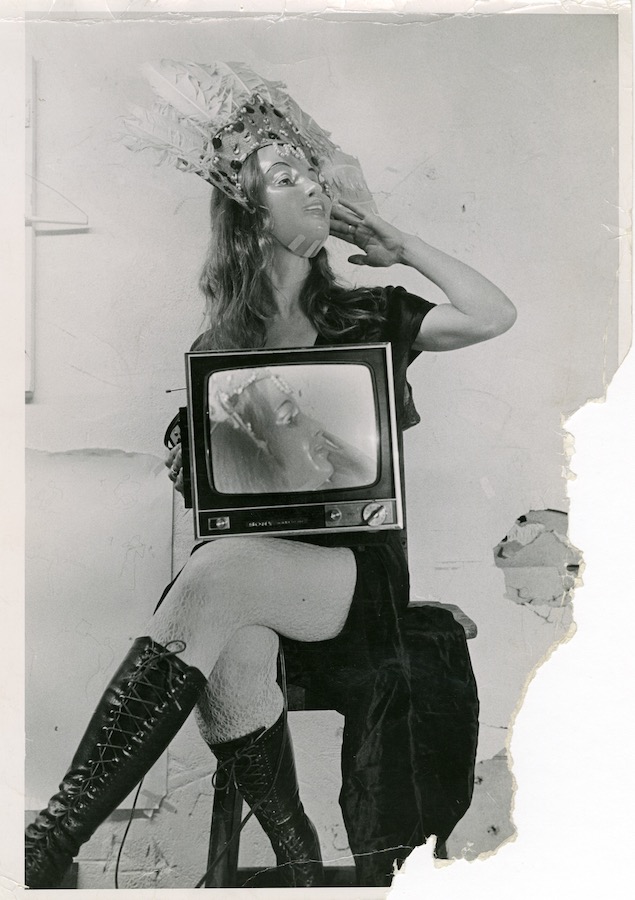 Jonas sits on a stool while crossing her legs and holding a TV monitor in her lap that shows a video of her face. Jonas is wearing a mask and a peacock-feathered headpiece.