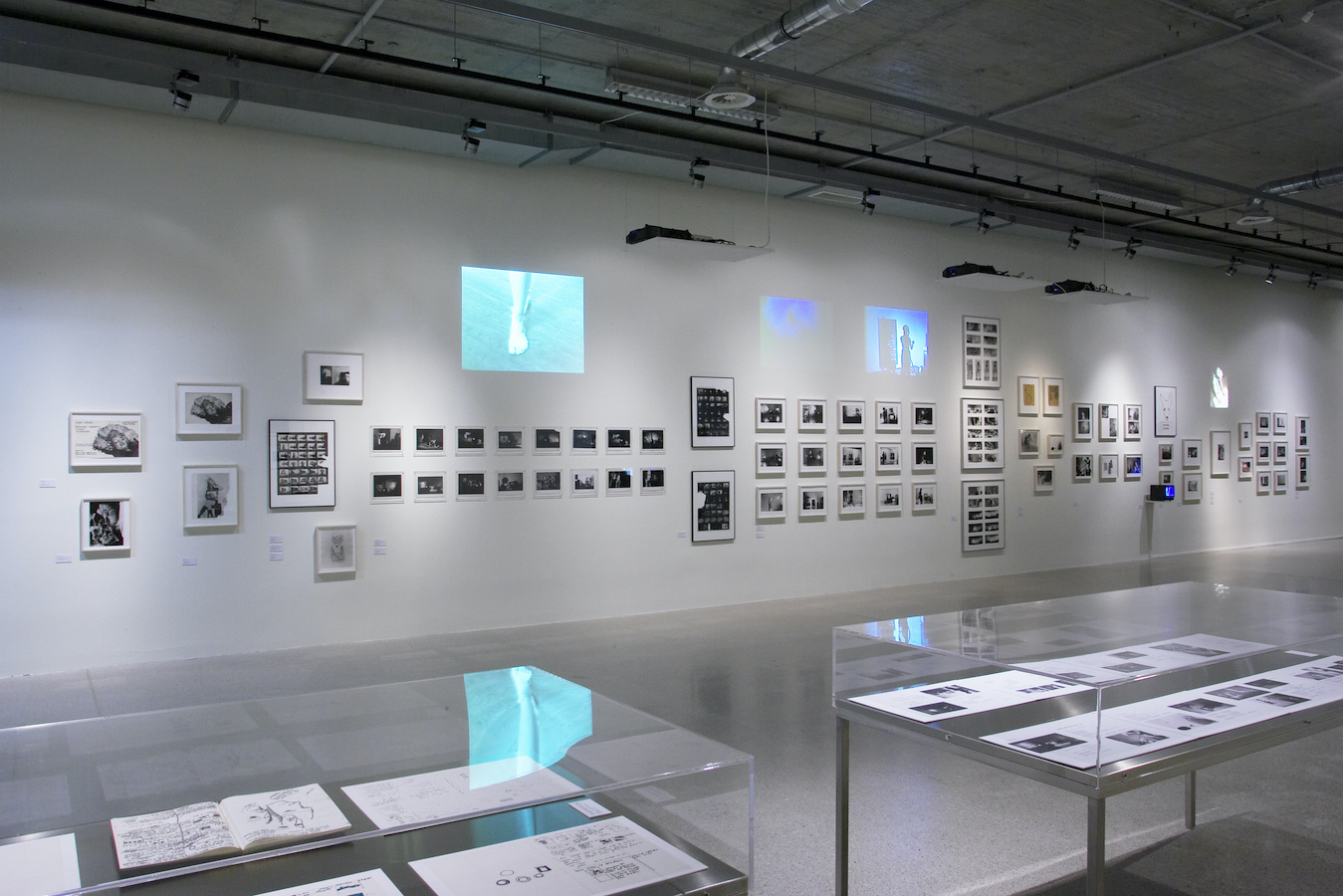 Two glass display cases hold various images and drawings. On the wall are multiple photographs, photographic contact sheets, and projections from Jonas’s performances.