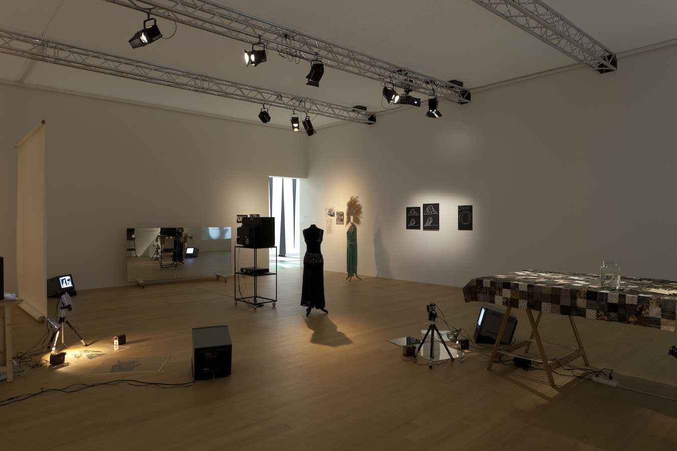 Various props from Jonas’s performance including: two mannequins wearing costumes, various drawings on the wall and floor, three TV monitors on the floor and one on a stand, a glass jar on top of a table draped in a cloth and two cameras on a tripod. To the left is a large mirror reflecting the room.