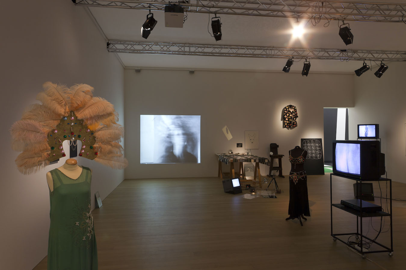 Various props from Jonas’s performance including: two mannequins wearing costumes, various drawings and a jacket hanging on the back wall, two TV monitors on stands and one on the floor, a table draped in a cloth, and a camera on a tripod placed above a mirror on the floor. There is a projection of Jonas’s performance on the wall.