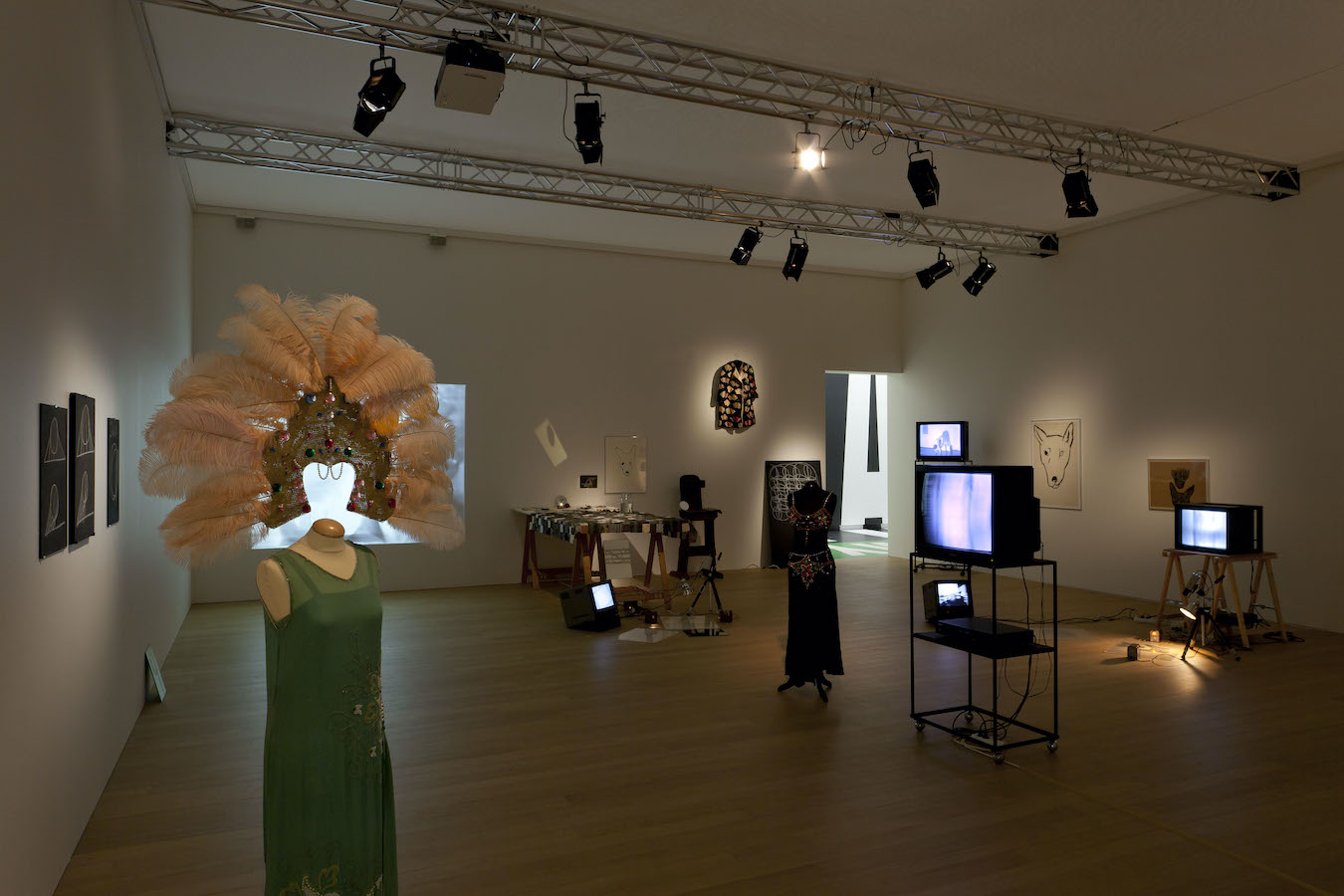 Various props from Jonas’s performance including: two mannequins wearing costumes, various drawings and a jacket hanging on the walls, three TV monitors on stands and one on the floor, a table draped in a cloth, and a camera on a tripod placed above a mirror on the floor. There is a projection of Jonas’s performance.