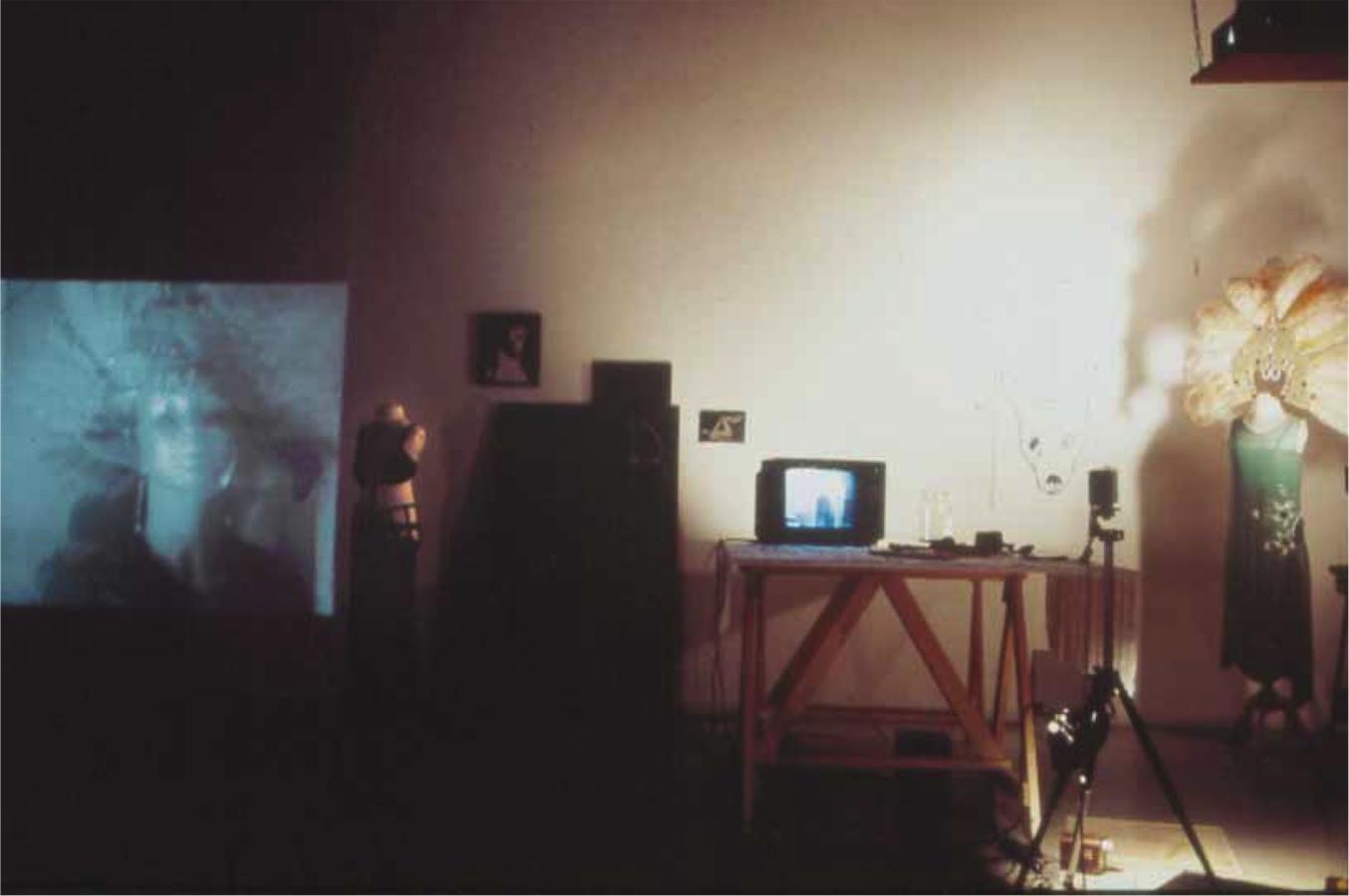 Various props from Jonas’s performance including: two mannequins wearing costumes, various drawings, a TV monitor placed on top of a table draped in a cloth, and a camera on a tripod with a light attached to it. To the left there is a projection of Jonas wearing a mask on the wall.