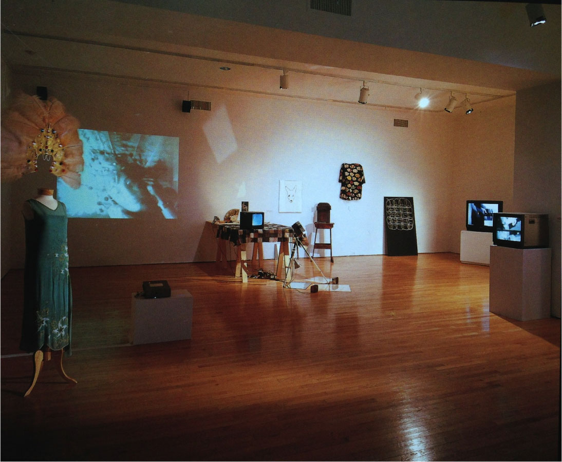 Various props from Jonas’s performance including: a mannequin wearing a dress and feathered headdress, various drawings on the floor and wall, three TV monitors: one is placed on top of a table draped in a cloth and the other two are placed on stands, and a camera on a tripod. There is a projection on the wall displaying a scene from Jonas’s performance.