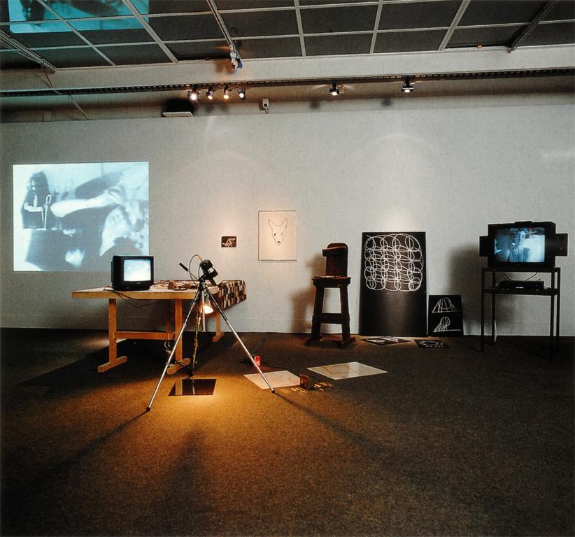 A camera on a tripod faces various props used for Jonas’s performance, a light is attached to the tripod and is pointed to the floor. There is a TV monitor placed on a table draped with a cloth; on the wall there is a projection to the far left and various drawings, including a dog’s head with four eyes, are in the middle. To the right of the drawings is a TV monitor on a stand displaying a scene from Jonas’s performance. On the floor are drawings and a mirror.