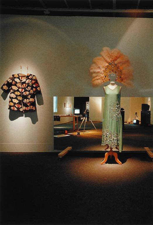 A mannequin draped in Jonas’s dress and peacock-feathered headpiece is placed in front of a mirror that is reflecting the rest of the room. To the left is a jacket hanging on the wall.