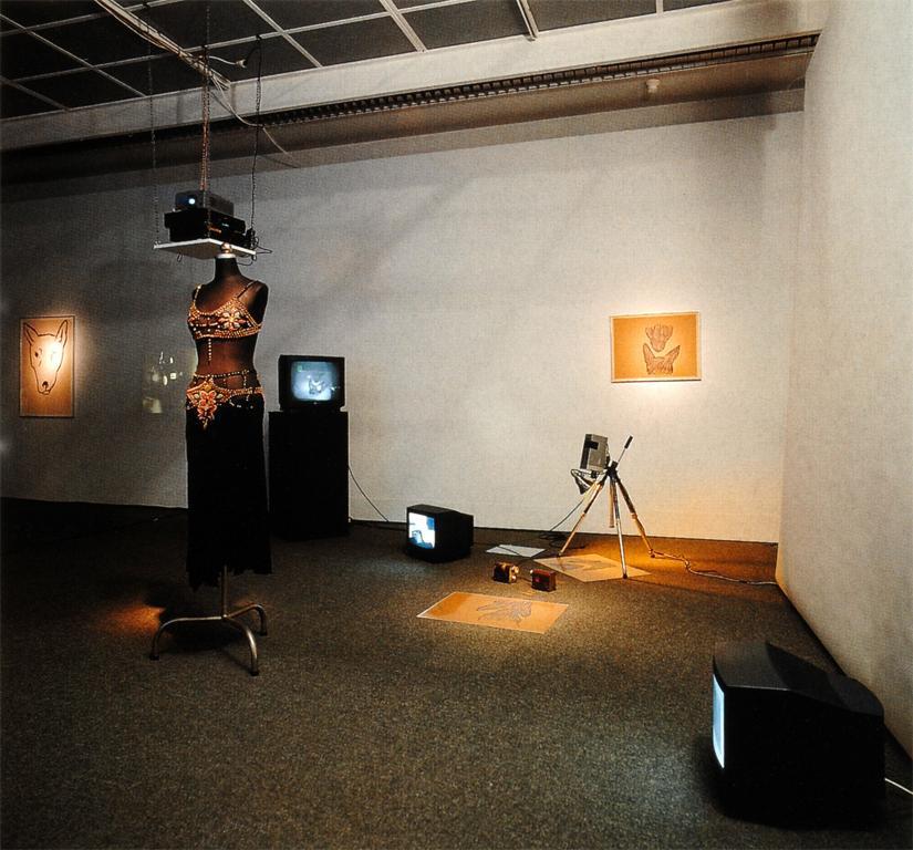 Various props from Jonas’s performance including: a mannequin wearing a costume, various drawings on the floor and walls, three TV monitors: two on the floor, and one on a stand, a projector hanging from the ceiling and a camera on a tripod. To the left is a drawing of a dog’s head.