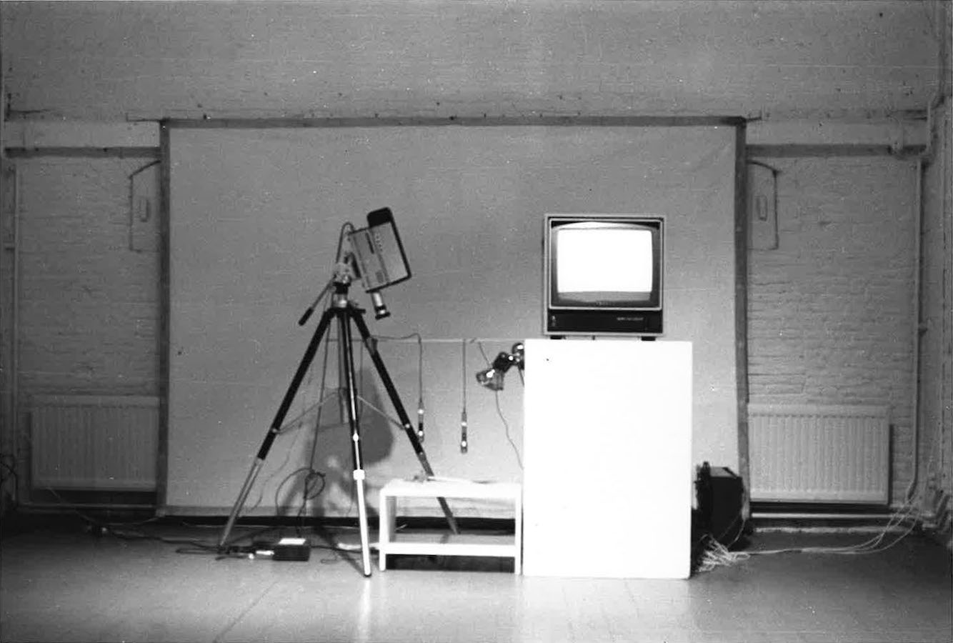 A camera on a tripod points down to two microphones hanging over a small white table. Next to the table is a video monitor on a white stand.