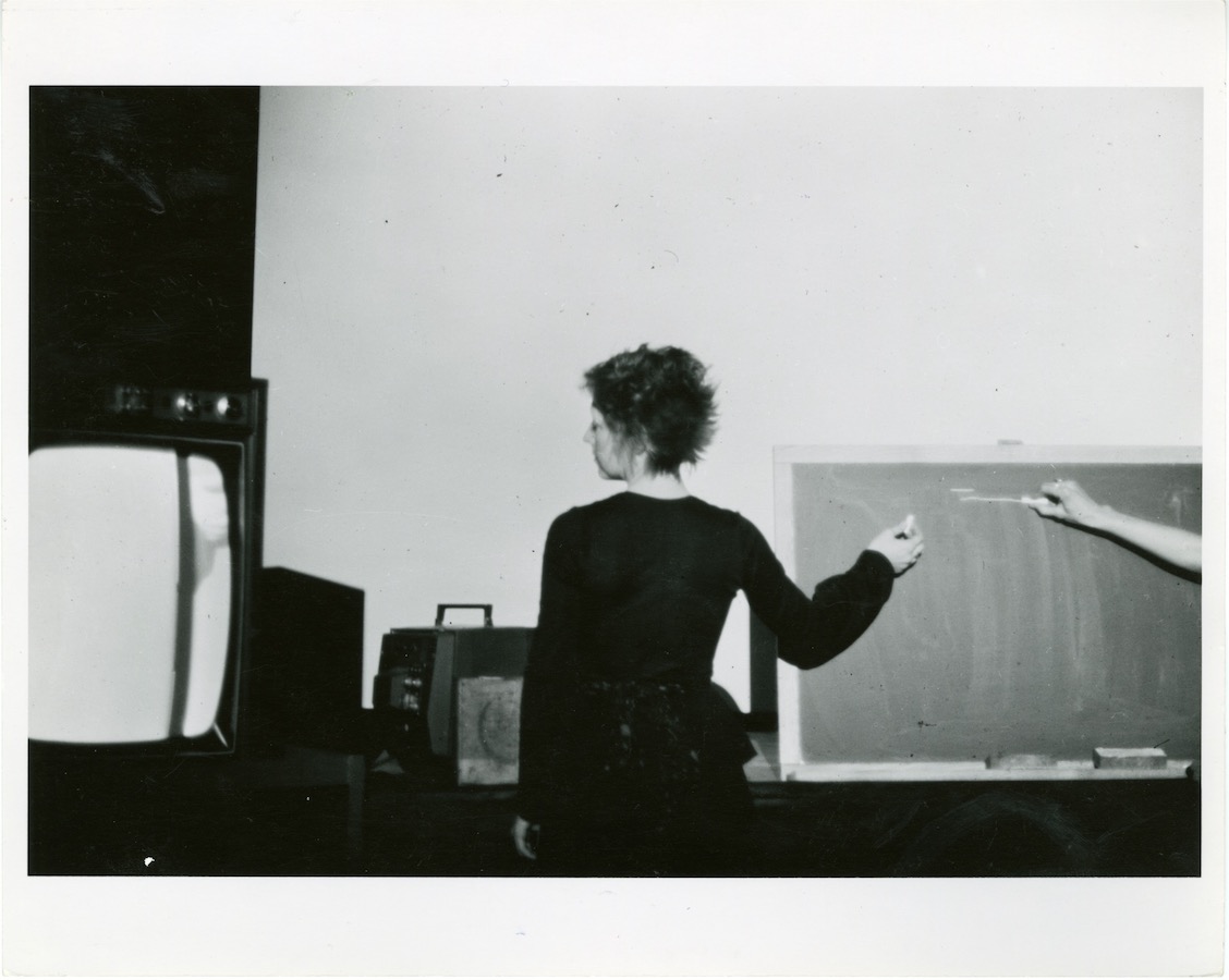 Jonas, with her back towards the camera, writes on a chalkboard to the right while looking at a TV monitor to the left. To the far right, another arm reaches in to write on the chalkboard.