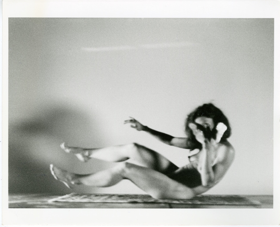 A naked Jonas sits on a rug with her feet hovering above the floor, her right arm stretched out in front and her left hand covers her face.