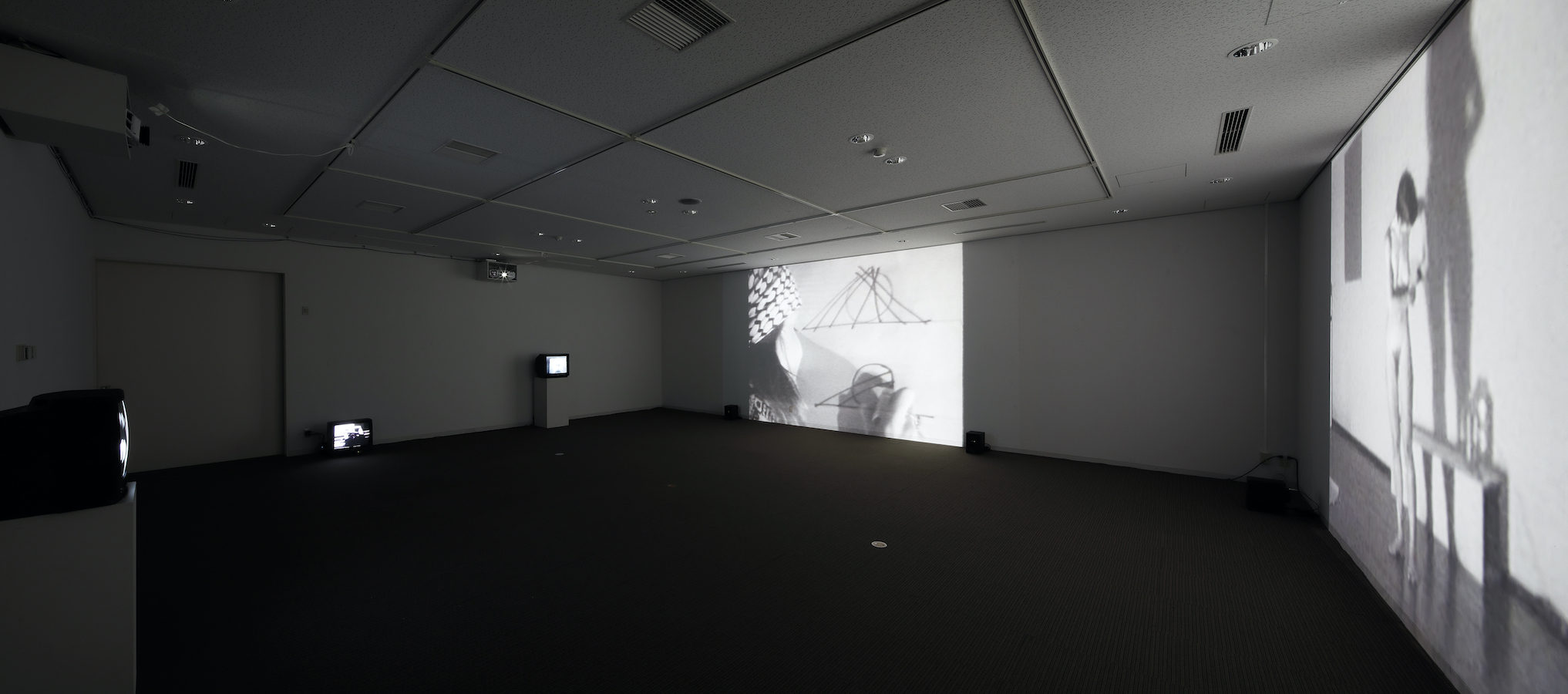 A dark room with two large projections on the walls and three TV monitors, two are on stands and one is on the floor.