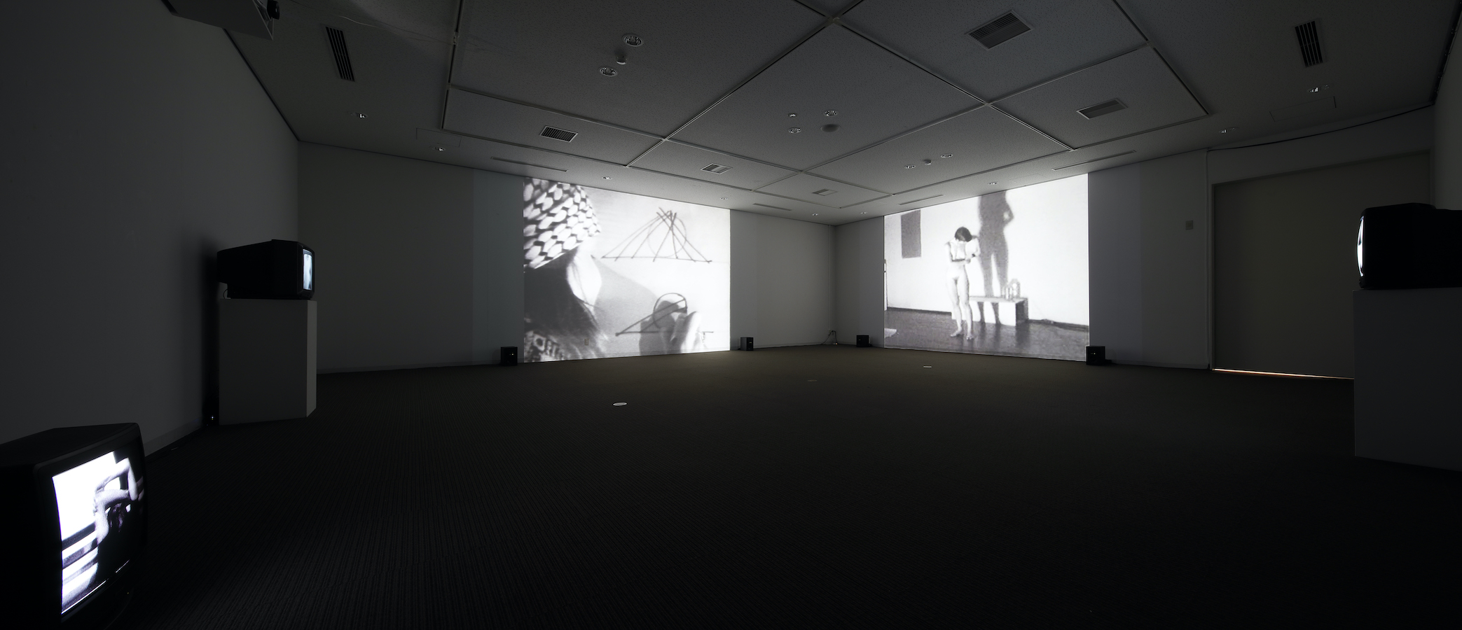 A dark room with two large projections on the walls and three TV monitors, two are on stands and one is on the floor.