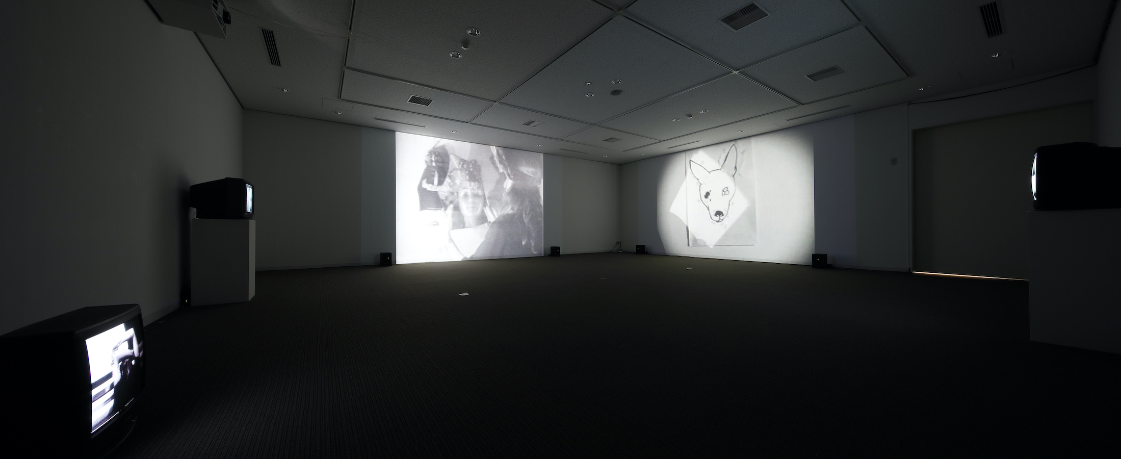 A dark room with two large projections on the walls and three TV monitors, two are on stands and one is on the floor.