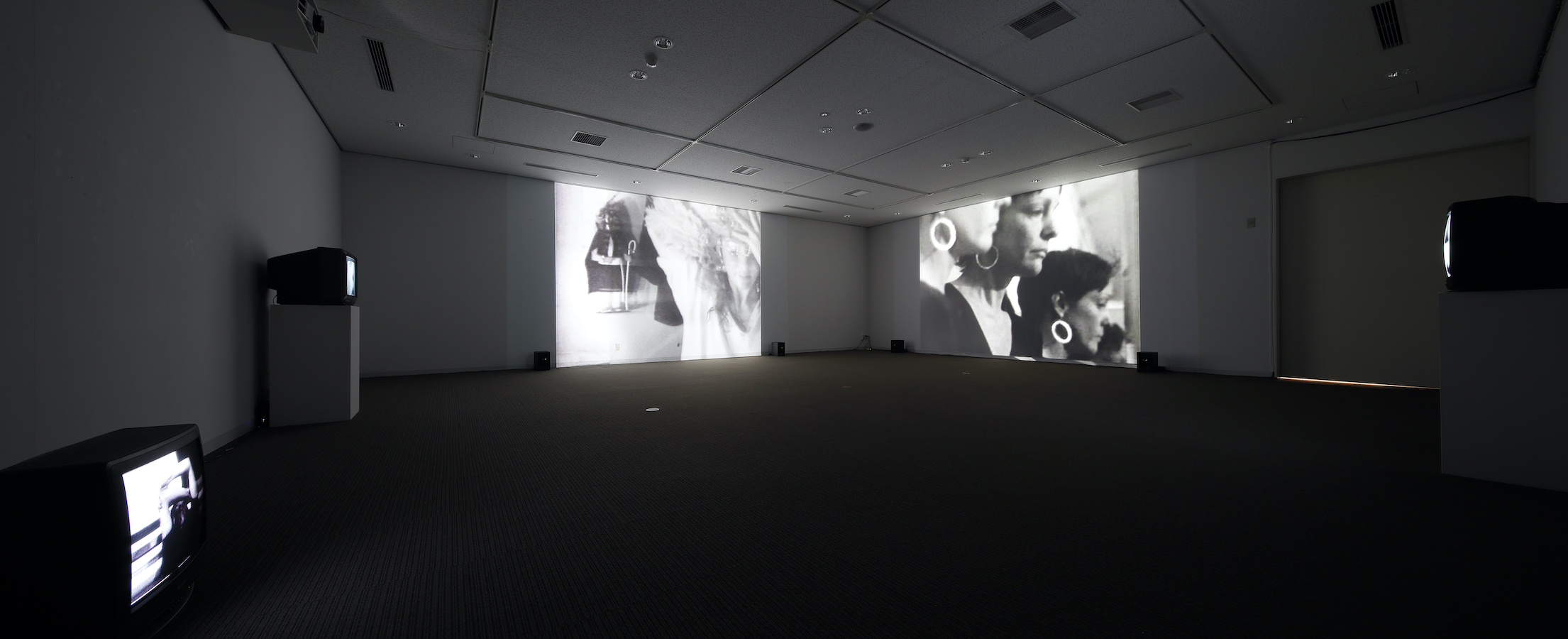 A dark room with two large projections on the walls and three TV monitors, two monitors are on stands and one is on the floor.