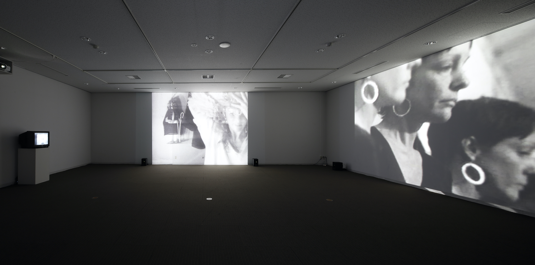 A dark room with two large projections on the walls and one TV monitor on a stand.