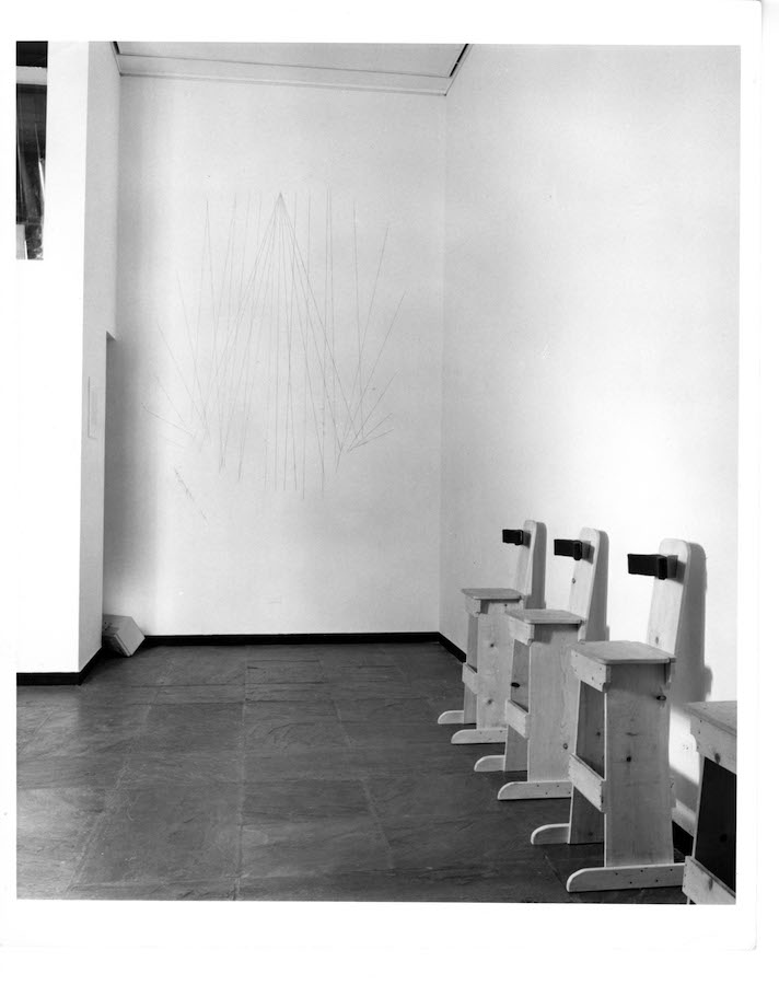 Wooden stools with backrests are lined up against the right wall. On the wall in the background is a line sketch of a cone.