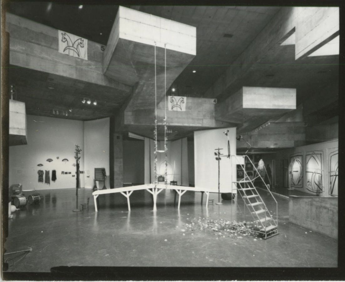 A swing hangs from the ceiling over a bridge placed in the middle of a room surrounded by various props; to the right of the bridge is a ladder.