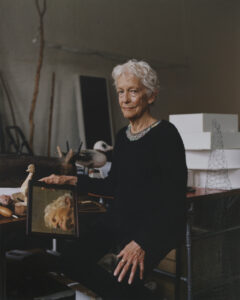 Jonas sitting on a stool surrounded by various props; in her right hand is a mirror reflecting her face.