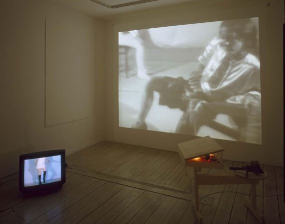 In the background is a projection of Jonas sitting. In the foreground to the left is a TV monitor on the floor displaying a body dancing and to the right is a two tiered table with a box on it.