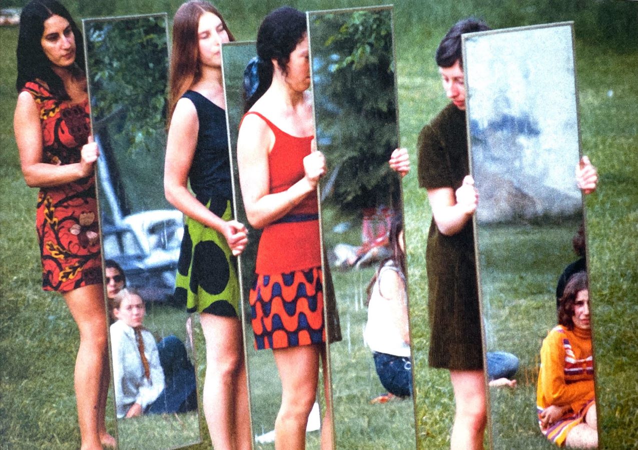 Four performers lined up in a row outside while holding full length mirrors that reflect the audience.