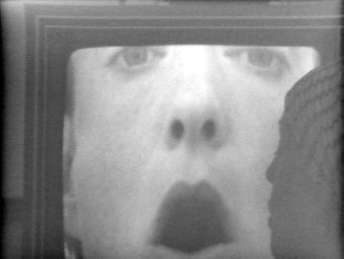 Video still of a closeup of Jonas’s face with her mouth open in an O-shape. To the right is a silhouette of another person’s face.