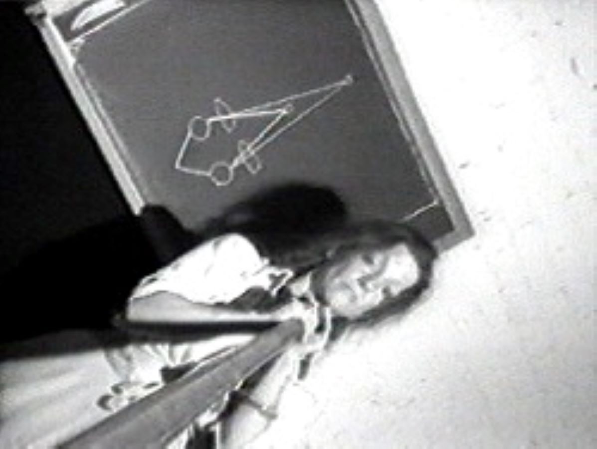 Video still of Jonas holding a large metal cone; in the background is a chalkboard with a drawing on it.