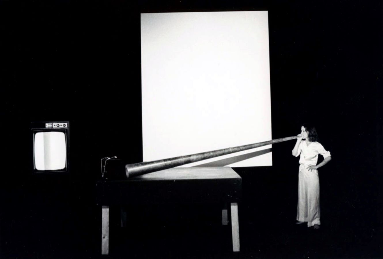 Jonas blowing into a large metal cone that is supported by a table. In the background is a blank screen and a TV monitor on a stand.