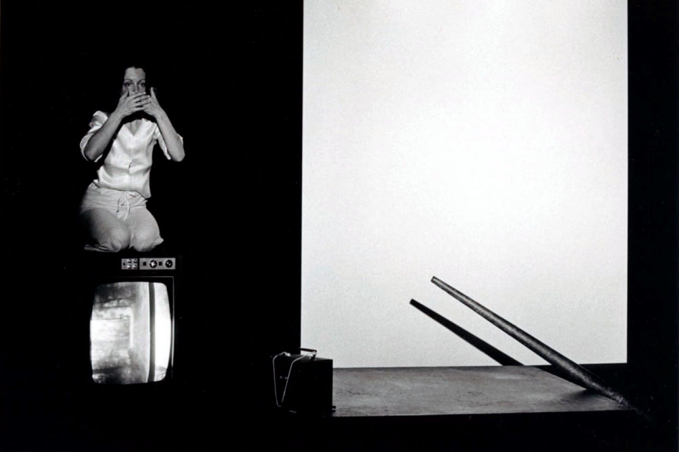 Jonas on top of a TV monitor with her hands over her mouth; to the right is a blank screen, a large metal cone, and a bag on top of a table.