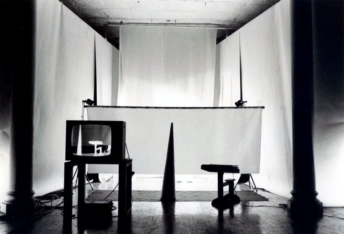 A cone and a stool are placed in front of white backdrops; in the foreground is a TV monitor on a stand displaying a negative image of the stool and cone.