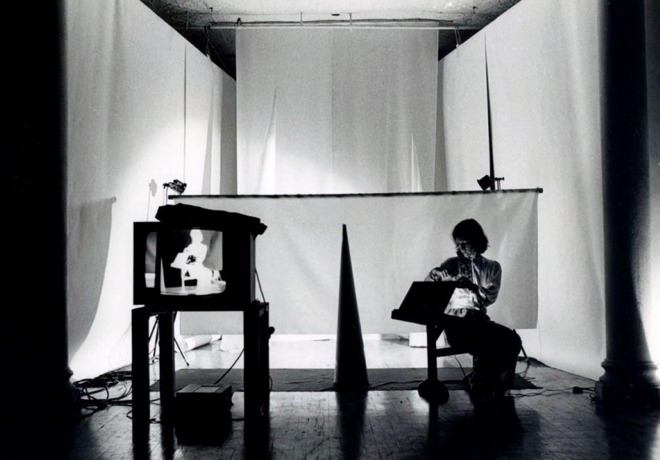 Jonas is sitting on a stool opening a box; in the background are white backdrops and a cone, in the foreground is a TV monitor on a stand displaying a negative image of Jonas