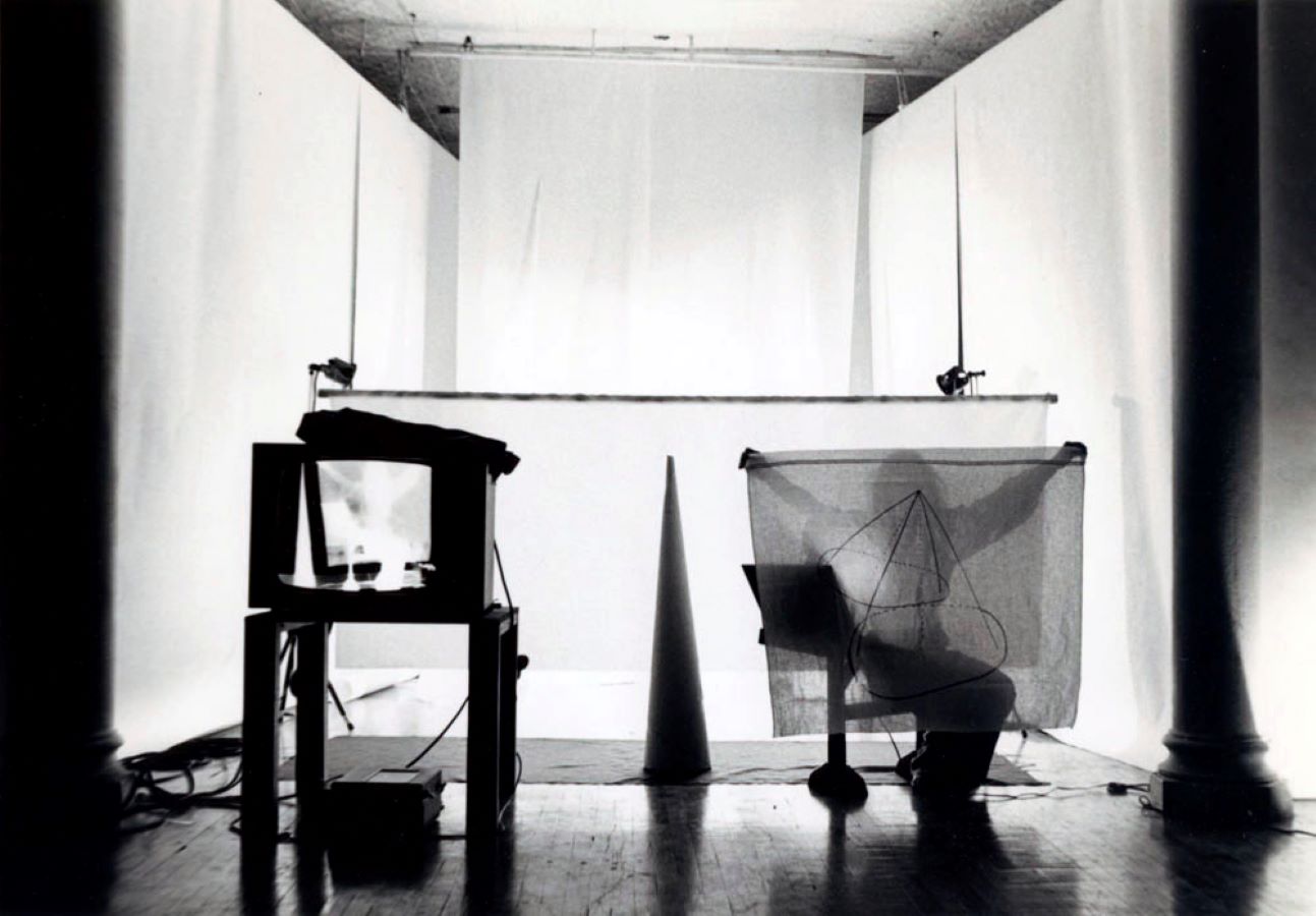 Jonas is sitting on a stool, holding up a sheet with a drawing on it; in the background are white backdrops and a cone, in the foreground is a TV monitor on a stand displaying a negative image of Jonas.